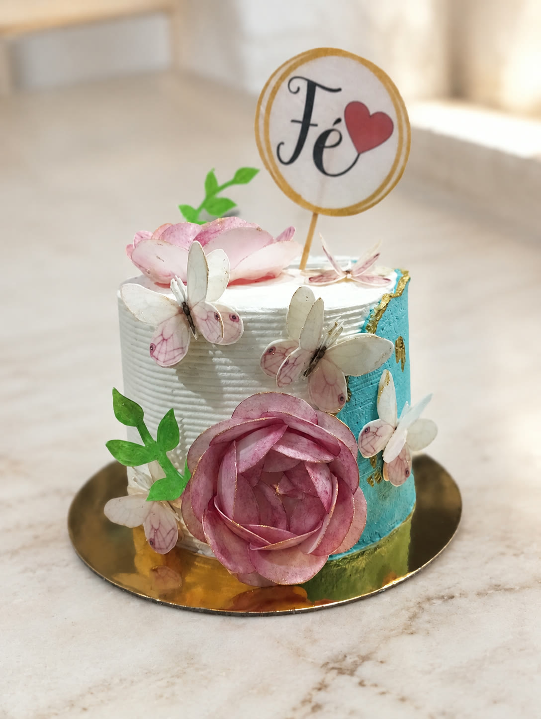 Bolo branco e azul decorado com flores, borboletas e a palavra Fé